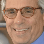 headshot of elderly white man with round brown glasses sitting in front of a bookshelf. picure is from the shoulders up. he's wearing a blue button up shirt and a red tie with a white circular design.