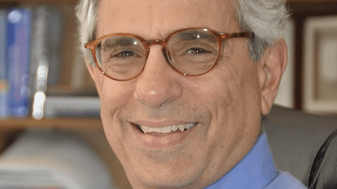 headshot of elderly white man with round brown glasses sitting in front of a bookshelf. picure is from the shoulders up. he's wearing a blue button up shirt and a red tie with a white circular design.