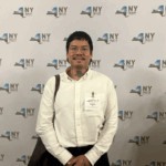 an Asian nonbinary person stands in front of a New York Health banner.