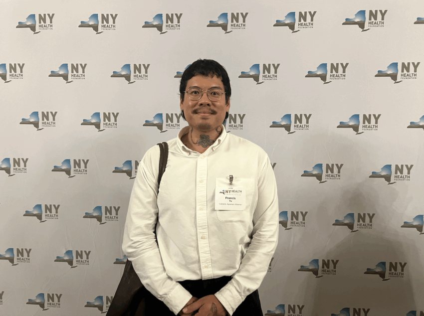 an Asian nonbinary person stands in front of a New York Health banner.
